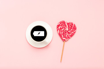 White cup with black coffee, pink heart-shaped lollipop, on a pink background. Sign a charged battery. Concept charge for the day. Flat lay, top view