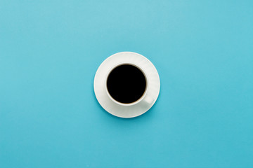 White cup with a saucer and black coffee on a blue background. Flat lay, top view