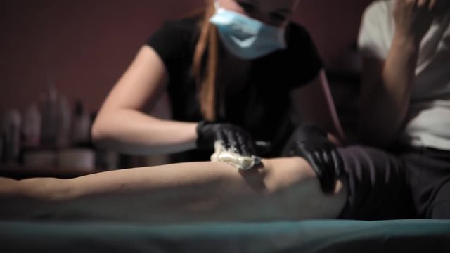 A sugaring is performed using the sugar paste from the legs of a young woman at a spa. The master in black gloves removes the hair in the direction of growth.