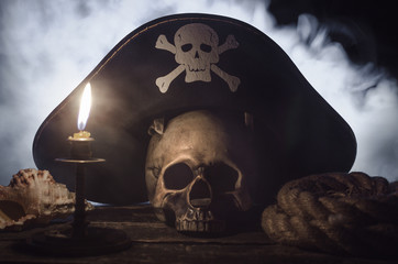 Obraz premium Ludzka czaszka z pirackim kapeluszem kapitana powyżej, płonąca świeca, muszla i lina cumownicza na drewnianym stole w mistycznym dymie.