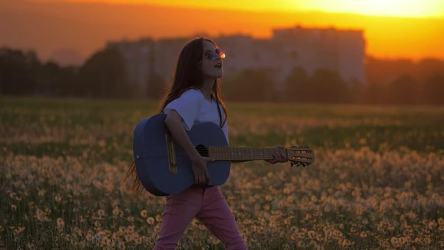 little girl teenager sunglasses during sunset, dancing and playing guitar standing in a field near the house. concept of celebration and entertainment