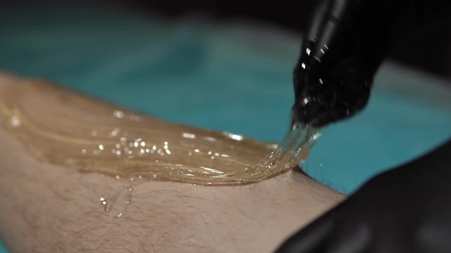 A sugaring is performed using the sugar paste from the legs of a young woman at a spa. The master in black gloves removes the hair in the direction of growth.