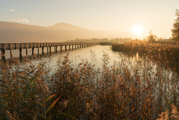 long wooden boardwalk pier over water in golden evening light with a mountain landscape silhouette in the background and golden marsh grass in the foreground