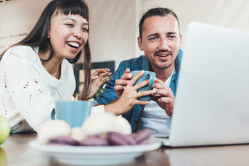 Beauty couple sitting together at the table with laptop watching movies, with cookies, fruits and hot drinks. Multiethnic young family