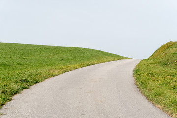 A horizontal view of an empty road leading into the blue sky along green grassy shoulders or embankments