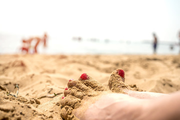 legs of a woman in the sand, nails made up red color, close-up, rest on the beach, blurred people on the seashore background