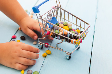 Hands of a child holding a shopping cart with colored sweets on a blue background