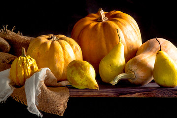 Large yellow pumpkins and pears lie on a rough cloth and gauze on a black wooden table and black background