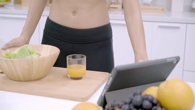 Young Asian woman making salad healthy food while using tablet for looking recipe and drinking orange juice in the kitchen, beautiful female preparing salad for fit body at home. Healthy food concept.