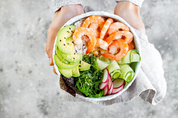 Girl holding shrimp poke bowl with seaweed, avocado, cucumber, radish, sesame seeds.
