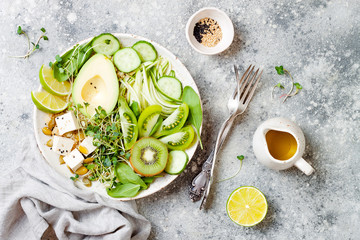 Vegan, detox Buddha bowl with quinoa, avocado, zucchini noodles, cucumber, tomato, lime, kiwi, tofu, spinach, micro greens, pepitas. Grey concrete background