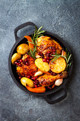 Roasted chicken legs with root vegetables, lemon, garlic, cranberry and rosemary on pan, on black slate stone background