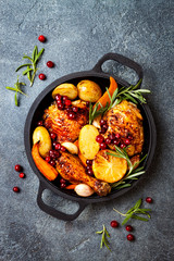 Roasted chicken legs with root vegetables, lemon, garlic, cranberry and rosemary on pan, on black slate stone background