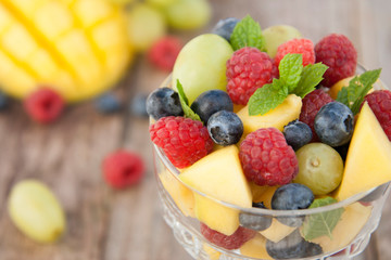 Delicious fruits salad in glass - raspberry, mango, blueberry, grapes, mint leaves. Wooden table, rustic background.