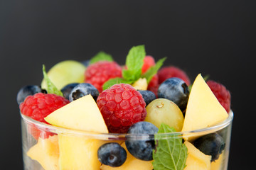 Close up, delicious fruits salad in glass - raspberry, mango, blueberry, grapes, mint leaves. Wooden table, rustic background.