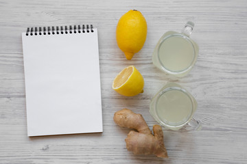 Glass jars of homemade ginger tea with lemon over white wooden background, top view. Blank notepad. Flat lay, overhead, from above.