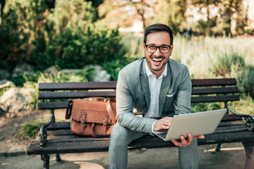 Fototapeta premium Portrait of a happy businessman using laptop outdoors. Sitting on the bench and looking at camera.