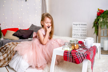 Obraz na płótnie Canvas Pretty little girl 4 years old in a pink dress. Child in the Christmas room with a bed, eating candy, chocolate, cookies and drinking tea, feeding teddy bears and laughing, good mood