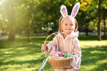 Cute little girl with bunny ears and basket of Easter eggs in park. Space for text