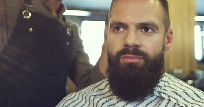 Portrait of the attractive caucasian man with a beard having his hair being cutted in the hairdressing. Close up.