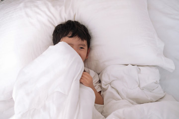 3 years old little cute Asian boy at home on the bed, kid lying playing and smiling on white bed with pillow and blanket, top view with copy space.
