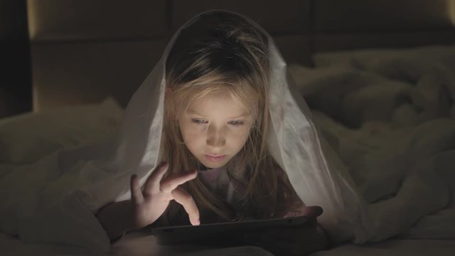 Teenage girl in bed playing a tablet in social internet in the dark light. Close up of little girl watching video on the digital tablet at night.