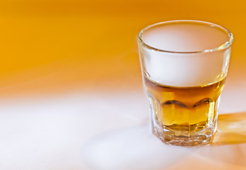 Ángulo levemente elevado de un pequeño vaso de whisky humeando aislado sobre un fondo naranja y blanco con luz lateral selectiva sobre él. 