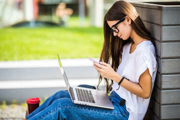 Pretty young woman sitting on bench in city on summer day and reading text message on cell phone while using silver laptop and drinking takeaway coffee