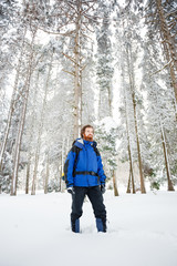 Fototapeta na wymiar Bearded young mountain climber standing in deep powder snow, surrounded by tall fir trees. Wide angle front view