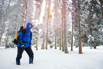 Fototapeta na wymiar Determined young male mountain climber hiking through deep powder snow in fir tree forest. Wearing heavy backpack with camping equipment. Active lifestyle in cold weather.