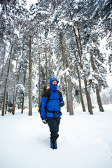Young male mountain climber wearing blue and black clothing hiking towards the camera through deep powder snow in fir tree forest. Wide angle frontal view. Active lifestyle in cold weather.