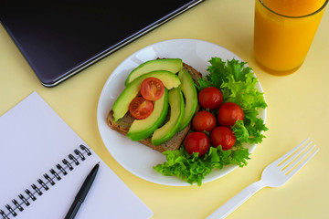 Healthy vegetarian snack with avocado, cherry tomatoes and lettuce leaves at workplace. Closed laptop, glass of juice, fork, notebook, pen on yellow background.