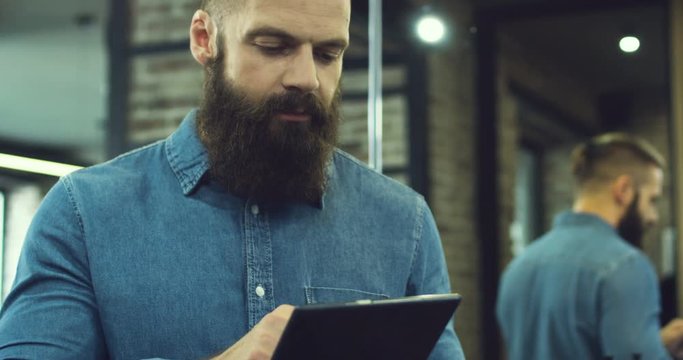 Close up of the handsome serious man with a beard in the blue jeans shirt scroling and taping on the tablet computer in the loft interior.