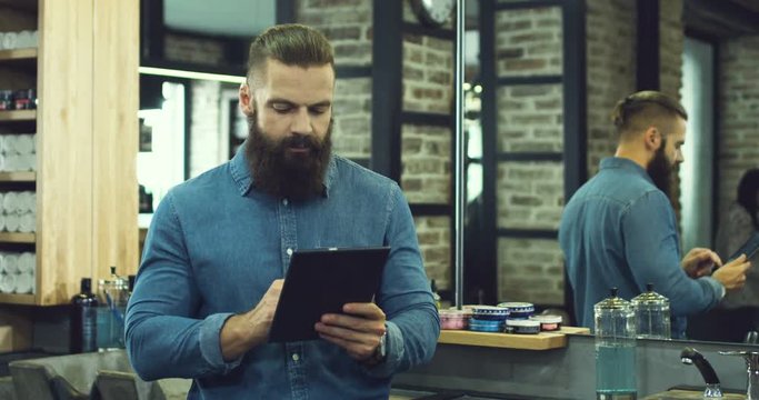 Portrait of the caucasian attractive serious man with a beard and in the blue jeans shirt typing ad taping on the tablet device in the barber shop.