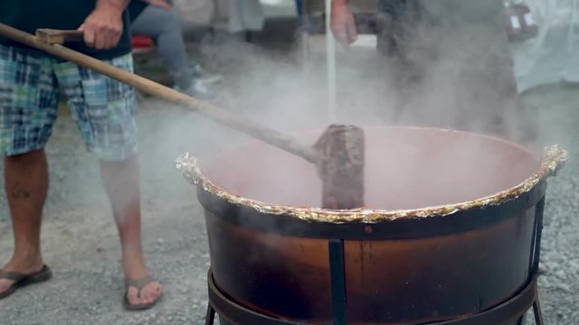 Very tight shot of stirring steaming apple butter in a large black cauldron outside.