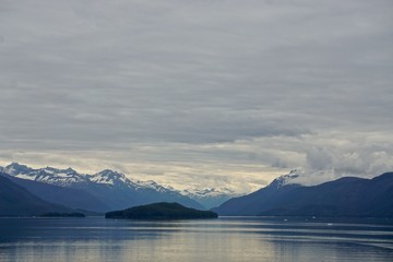 Obraz na płótnie Canvas Endicott Arm, Alaska, USA: Snow-capped mountains behind a small island, under a cloudy sky in the Endicott Arm, a fjord in the Pacific Northwest.