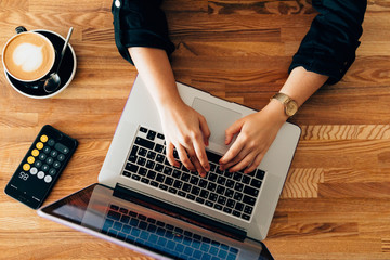 The girl works at the computer and drinks coffee from a cup in a cozy and bright cafe