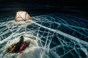 Fire on ice at night. Campground on ice. Tent stands next to bonfire. Lake Baikal. Nearby there is car. Shelter tent and ice are illuminated from the inside. Beautiful bonfire on real cracked ice
