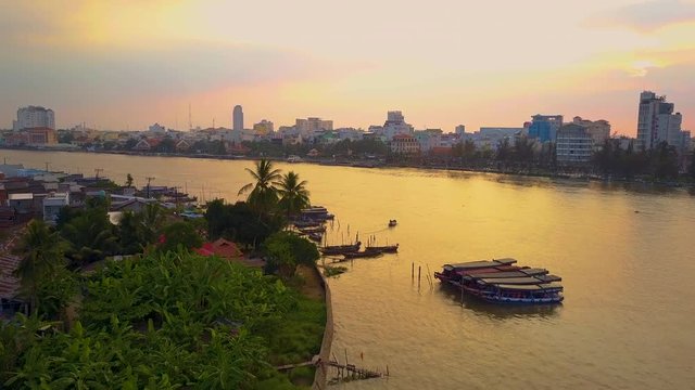 AERIAL: Small local boat cruises across the tranquil river to a large Vietnamese town at sunset. Flying behind a wooden boat and towards the idyllic waterfront city on a beautiful golden lit evening.
