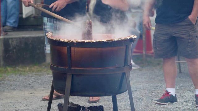 Closeup of someone stirring steaming apple butter in a large wood fired cauldron outside.