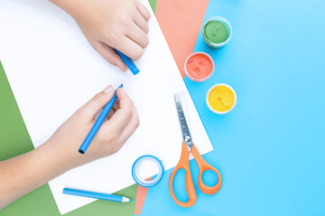 Color paper in blue, pink and green, paint, scissors and hands of a child drawing with blue pen top view flat lay, copy space