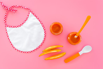 Baby food concept. Pumpkin puree in bowl near bib, pumpkin slices, spoon on pink background top view