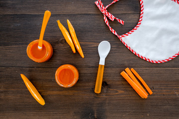 Healthy food for small babies. Carrot puree in bowl near bib, carrot slices, spoon on dark wooden...