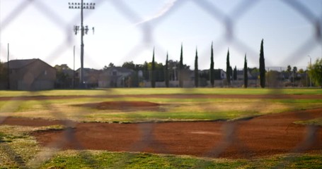 Slow slide left looking out on an empty green baseball field diamond from behind a chain link fence in a local park at sunrise.