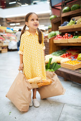 Cheerful excited girl in yellow checkered dress looking up and carrying full shopping bags in farmers market