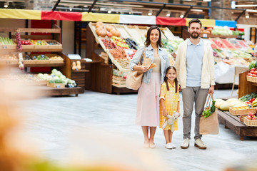Positive beautiful young family in casual clothing standing in farmers market and holding shopping bags while looking at camera