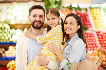 Cheerful friendly young family embracing and standing organic food store while looking at camera