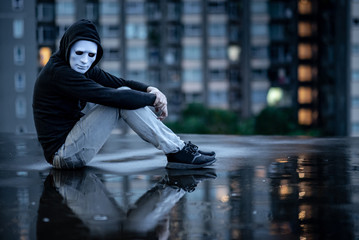 Reflection of mystery hoodie man in white mask hugging his knees sitting in the rain on rooftop of abandoned building. Bipolar disorder or Major depressive disorder. Depression concept