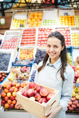 Happy modern attractive young lady entrepreneur in apron holding wooden box of red apples and smiling at camera