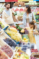 Positive beautiful young family choosing fresh fruits and vegetables while walking together in farmers market